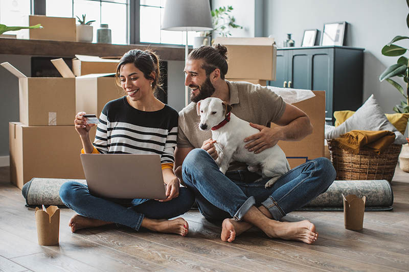 Young couple moving in new house. They are happy and using laptop to online shop with a dog on his lap.