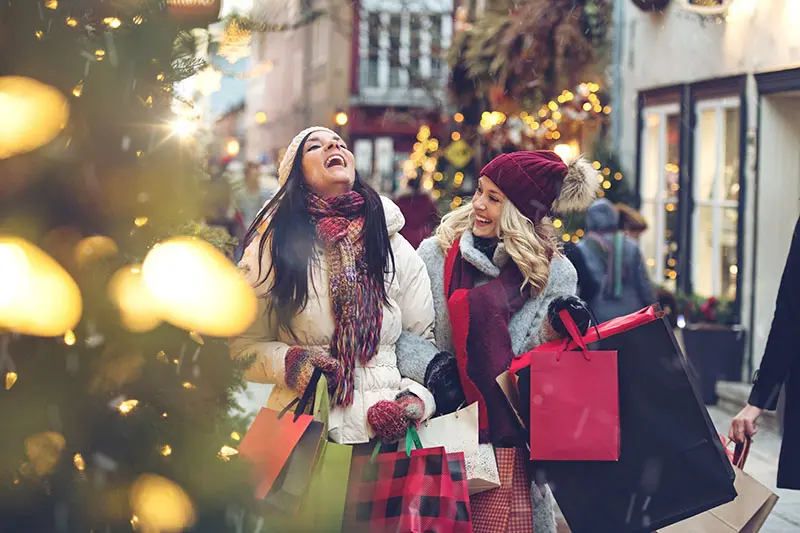 Two woman who are friends wearing winter coats while carrying lots of shopping bags are laughing in front of a Christmas Tree