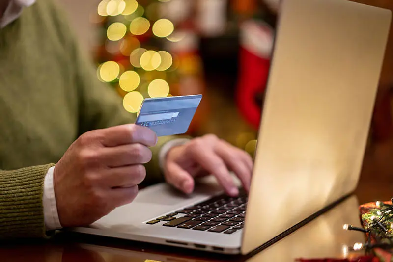 A man with a green sweater is holding his credit card while using his MacBook to shop online. It is the holiday season with light twinkle s and stockings in the background.