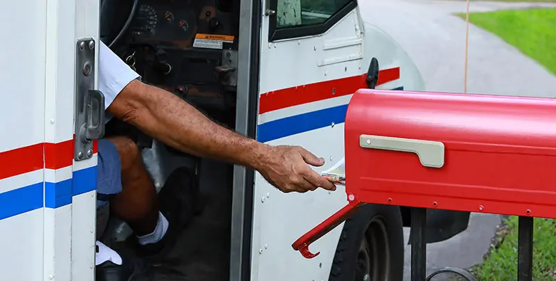USPS delivery driver in a vehicle delivering mail to a red mailbox on a residential street